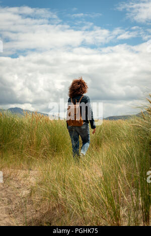 Jeune belle femme avec des cheveux bouclés rouges portant une veste bleue décontractée et un Jean bleu marchant sur la dune de sable côtière à travers l'herbe de marram en été Banque D'Images