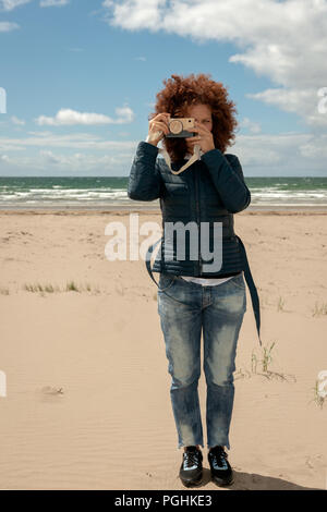 Belle jeune femme aux cheveux rouge bouclés tenant un appareil photo en bois sur la plage de sable vide de Strand, en Irlande Banque D'Images