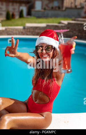 Belle jeune femme près de la piscine dans la région de Santa Claus hat célébrer Noël et Nouvel An en pays chaud avec un verre de cocktail. Banque D'Images