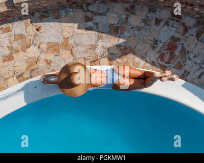 Belle jeune femme bénéficiant d'une piscine. Elle est couchée de soleil, avait couvert avec un énorme chapeau de paille, près de l'eau bleu clair. Banque D'Images