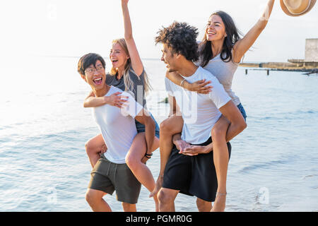 Groupe d'amis, marcher le long de la plage, avec des hommes en donnant piggyback ride à leur petite amie. Heureux les jeunes amis profiter d'une journée à beach Banque D'Images