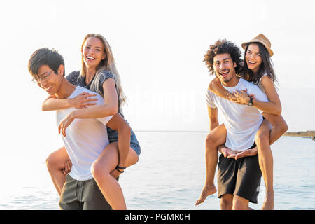 Groupe d'amis, marcher le long de la plage, avec des hommes en donnant piggyback ride à leur petite amie. Heureux les jeunes amis profiter d'une journée à beach Banque D'Images