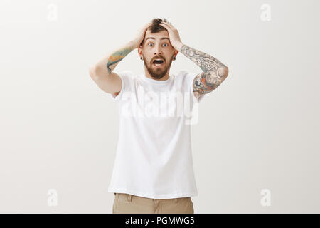 Guy entendu horrible nouvelle, étant très choqué. Portrait d'homme barbu emotive étourdis nerveux en t-shirt blanc, tenant les mains sur la tête et crier de peur, debout sur fond gris Banque D'Images