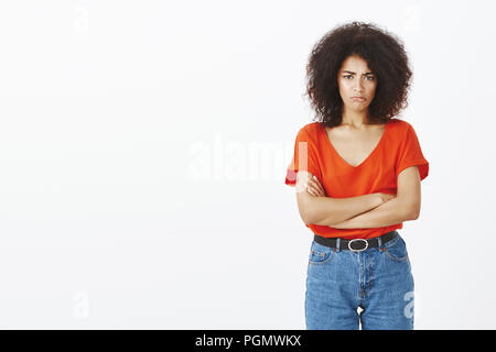 Studio shot of attractive offensé sombre femme aux cheveux bouclés avec la peau foncée, bouder tenant les mains croisées sur la poitrine et les sourcils, d'être mécontent ou seule, debout sur un mur gris insulté Banque D'Images