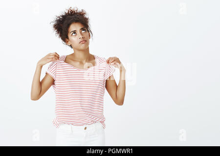 Contrarié de bouder la peau sombre femme avec afro coiffure, jusqu'à froncer et de mécontents des émotions, tirant sur t-shirt tout en sensation de chaleur, d'être malheureux en été de marche jour sur mur gris Banque D'Images