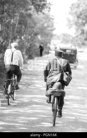 Vue nostalgique en noir et blanc, village de tramway de Crich, événement d'été des années 1940. Vue arrière de deux hommes qui font du vélo et suivent des véhicules d'époque au soleil. Banque D'Images