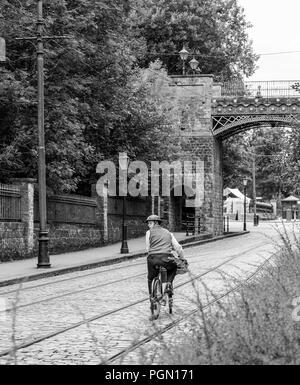 Vue arrière nostalgique, en noir et blanc, d'un cycliste solitaire des années 1940, dans un casque en étain, longeant une rue pavée déserte qui donne sur l'arrière. Banque D'Images