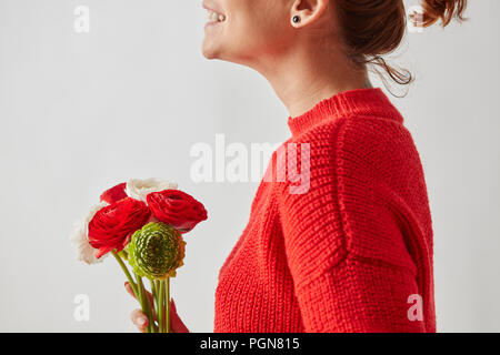 Happy girl in a red chandail tricoté avec un magnifique bouquet de fleurs aux couleurs vives Ranunculus autour d'un arrière-plan gris avec copie espace. Banque D'Images