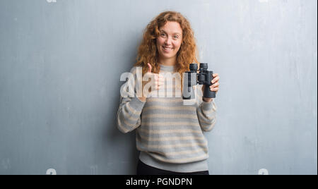 Jeune femme rousse sur mur gris grunge binoculars heureux avec un grand sourire faisant signe ok, pouce vers le haut avec les doigts, signe d'excellentes Banque D'Images