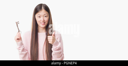 Jeune femme de l'agent de l'état réel chinois sur fond isolé holding vintage key heureux avec un grand sourire faisant signe ok, pouce vers le haut avec les doigts, excellent Banque D'Images