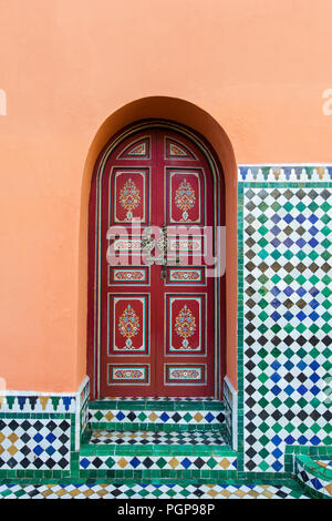 Extérieur de porte voûtée marocain, peints en rouge avec motifs floraux, encadré dans un mur de plâtre orange décoré de mosaïque. Marrakech, Maroc Banque D'Images