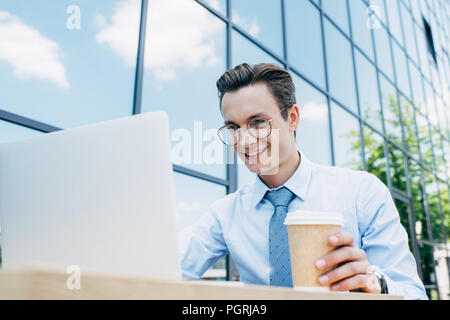 Handsome smiling businessman using laptop and lunettes holding paper cup à l'extérieur du bâtiment moderne Banque D'Images