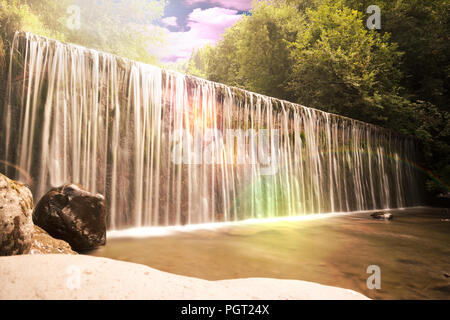 L'eau qui coule sur un rebord ou mur de barrage dans une cascade cascade pittoresque ou avec feuillage vert luxuriant et sun flare avec rainbow au coucher du soleil Banque D'Images