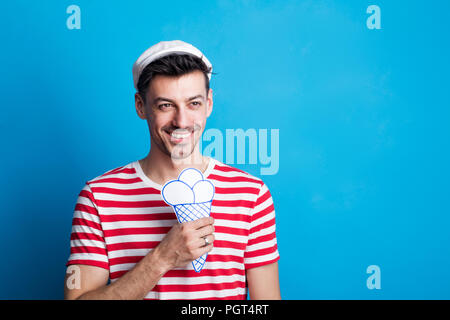 Portrait d'un jeune homme dans un studio avec des lunettes de soleil, d'un capuchon tenant une glace papier sur un fond bleu. Copier l'espace. Maison de concept. Banque D'Images