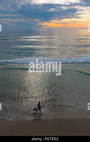 Sunset seascape, orange, bleu ciel, jaune, vert aqua marine avec la mer, les vagues blanches dans le matériel roulant, surfeur au crépuscule balade dans le surf, transportant son livre blanc Banque D'Images