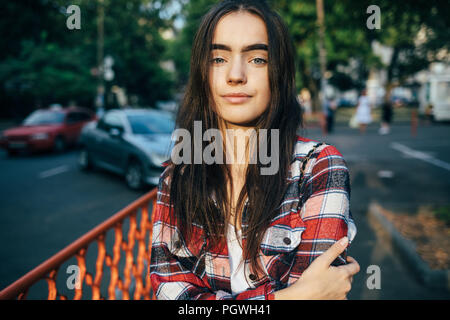 Teen fille avec de longs cheveux noirs wearing plaid shirt regarde la caméra et sourit. Portrait of happy young woman standing in city bénéficiant de Banque D'Images