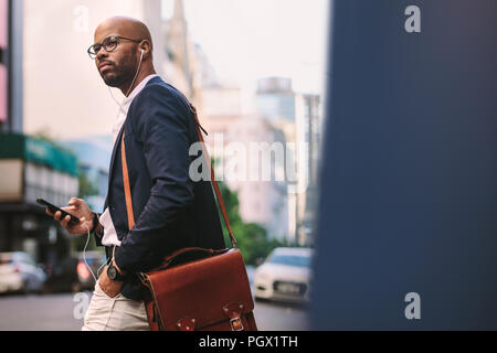 African businessman avec sac en marchant dans la rue avec un téléphone mobile. Handsome businessman listening music de smart phone en marchant sur stre ville Banque D'Images