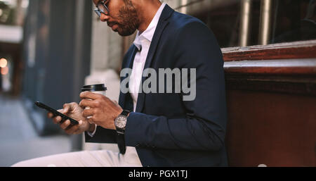 African businessman assis dehors avec du café et à l'aide de téléphone mobile. Man in suit de détente en plein air par la route des textos sur son portable et drinkin Banque D'Images