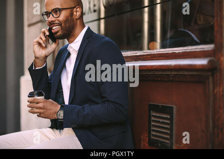 Heureux homme africain de détente en plein air avec café et conversation sur téléphone mobile. Jeune homme en costume assis à l'extérieur et à parler au téléphone. Banque D'Images