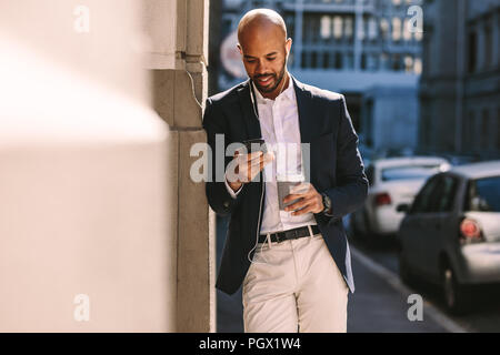 Handsome businessman leaning on un mur tout en se tenant à l'extérieur et l'utilisation de smartphone. Man wearing earphones en regardant son téléphone mobile. Banque D'Images