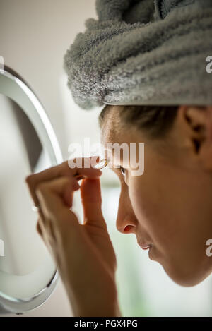 Femme avec ses cheveux enroulée dans une serviette avant de Pise plucking her eyebrows attentivement dans le miroir. Banque D'Images