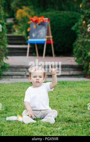 Drôle de Portrait Portrait blanc tout-petit enfant kid boy sitting on grass terre à l'extérieur en été automne parc en dessin avec chevalet books montrant roc Banque D'Images