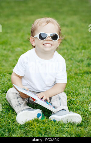 Portrait of smiling rire funny cute adorable bambin garçon caucasien enfant blanc avec des cheveux blonds en blanc T-shirt et lunettes de soleil assis sur vert gr Banque D'Images