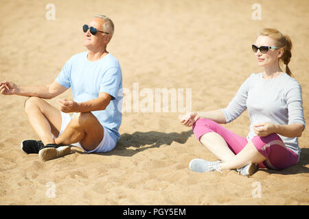 Couple contemporain activewear et lunettes assis sur le sable en posture de lotus après entraînement Banque D'Images