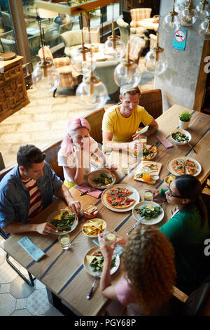 Cinq jeunes amis assis par de grandes tables en bois et profiter a servi de la nourriture et de parler dans un café ou restaurant Banque D'Images