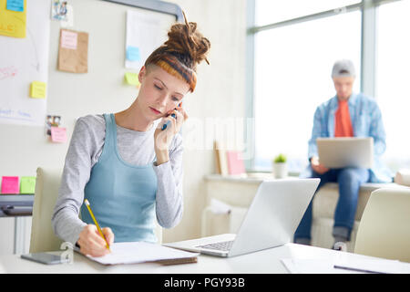 Hipster girl concentré grave avec dreads talking on mobile phone et prendre des notes en bloc tout en travaillant dans un bureau moderne Banque D'Images