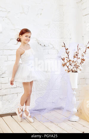 Young ballerina girl se prépare pour un ballet. Peu de prima ballet. Jeune fille dans une robe de bal blanche et Pointe près de la fenêtre, belle rouge h Banque D'Images