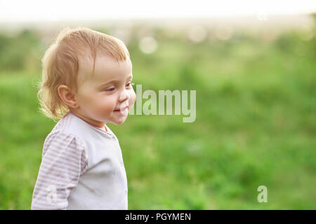 Portrait of smiling petit enfant marche sur champ vert. Girl à heureux, positif, portant une chemise blanche, ayant de grands yeux verts, des cheveux blonds courts, Banque D'Images