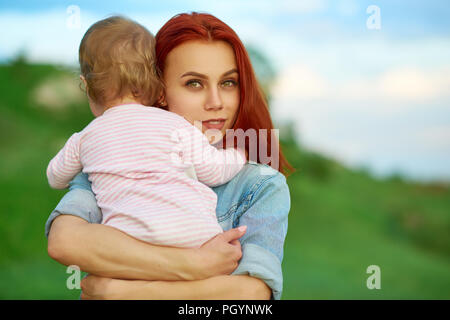 Jeune mère serrant mignon petit enfant posant sur champ vert. Woman looking at camera, ayant les caractéristiques du visage magnifique, de grands yeux, porter un jean bleu shi Banque D'Images