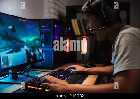 Portrait of asian angry gamer boy à jouer à des jeux vidéo sur ordinateur dans la pièce sombre portant des écouteurs et à l'aide clavier coloré rétroéclairé Banque D'Images