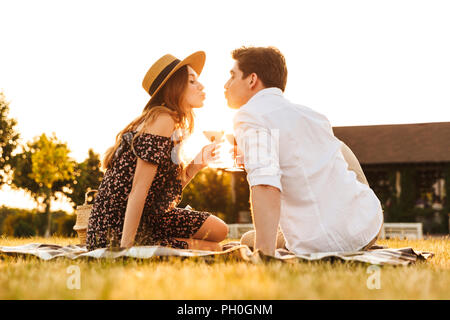 Image de jeune couple aimant assis par dating outdors on picnic holding verres de vin s'embrassant. Banque D'Images