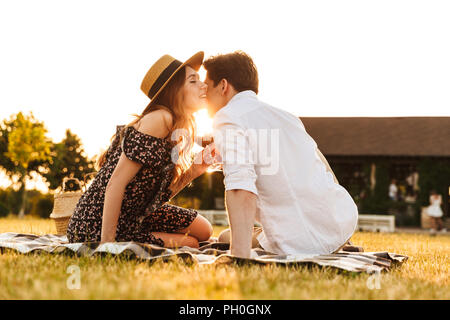 Image de jeune couple aimant assis par dating outdors on picnic holding verres de vin s'embrassant. Banque D'Images