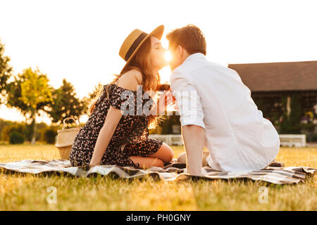 Image de jeune couple aimant assis par dating outdors on picnic holding verres de vin s'embrassant. Banque D'Images