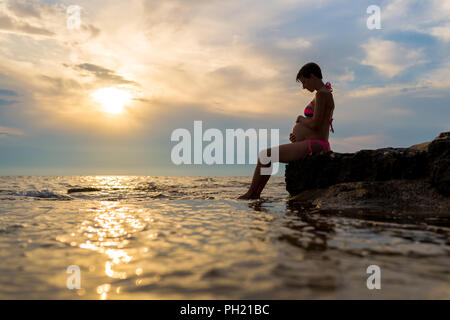 Femme enceinte en maillot assis de profil, berçant son ventre sur un rocher au bord de la mer au coucher du soleil avec le soleil jette une golden beam partout e Banque D'Images