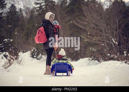 Image style vintage rétro d'une jeune mère tirant son enfant à l'hiver la neige sur un traîneau en tournant et en lui souriant dans une forêt de pins de montagne. Banque D'Images