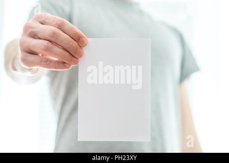 Homme dans un t-shirt gris tenant une feuille de papier blanc vierge dans sa main tendue avec copyspace pour votre texte ou une publicité. Banque D'Images