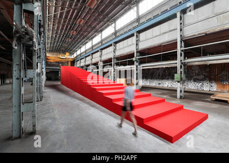 L'Allemagne, Leipzig. Août 24, 2018. Une femme marche le long d'une installation semi-finis par Victor Freso, un immense escalier rouge. Le Festival d'Art Urbain 'Monumenta' ouvre le 1er septembre dans l'usine vide de l'ex-Pittlerwerke salles. La Monumenta présente le graffiti, le street art et l'art contemporain autour de 6 000 mètres carrés jusqu'au 13 octobre. Crédit : Jan Woitas/dpa-Zentralbild/dpa/Alamy Live News Banque D'Images