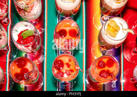 Belle desserts faits en couches de crème, baies et physalis dans les hautes wineglasses avec une cuillères debout dans une ligne sur une tablecl multicolore dynamique Banque D'Images