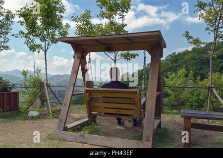 Vue arrière de l'homme blond assis sur balançoire en bois et en regardant le paysage magnifique Banque D'Images