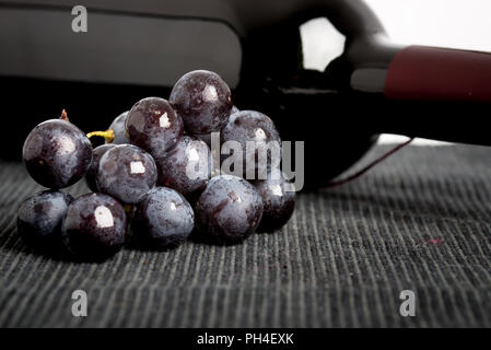 Tas de raisins noirs frais et qui contient une bouteille de vin rouge couché sur le côté sur un textile gris avec copyspace conceptual de la vinification, de l'œnologie Banque D'Images