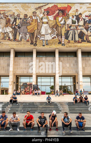 Les jeunes se détendre sur les étapes de la Musée Historique National, ci-dessous, l'ère communiste, mosaïque murale, dans la place Skanderbeg, Tirana, Albanie, Banque D'Images