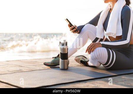 Photo recadrée de jeune femme de remise en forme en plein air sur la plage ont un reste assis avec une serviette et de l'eau à l'aide de téléphone mobile. Banque D'Images