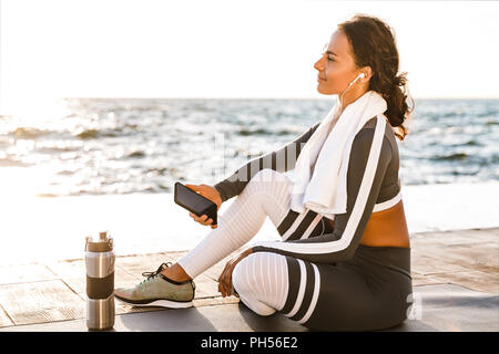 Image de jeune femme souriante de remise en forme en plein air sur la plage ont un reste assis avec serviette à côté d'écouter la musique avec vos écouteurs. Banque D'Images