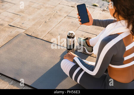 Photo recadrée de jeune femme de remise en forme en plein air sur la plage ont un reste assis avec une serviette et de l'eau à l'aide de téléphone mobile. Banque D'Images