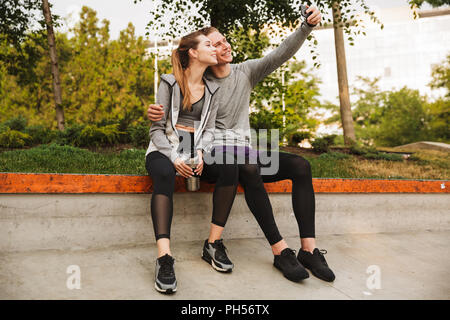 Image de young couple homme et femme 20s en survêtements assis sur le parapet en Green Park et après entraînement selfies ou courir sur d'été ensoleillé Banque D'Images