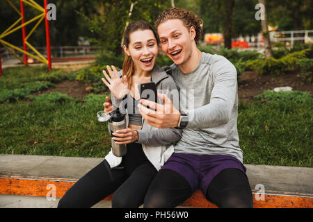 Photo de cheerful couple sport homme et femme 20s dans les vêtements de sport assis sur parapet dans Green Park et après entraînement selfies ou courir sur été ensoleillé Banque D'Images
